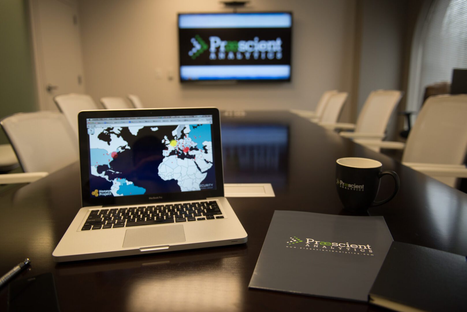 Conference room with a laptop displaying a world map, a "Prescient" branded mug, and a folder on a table. A screen in the background shows the Prescient logo.