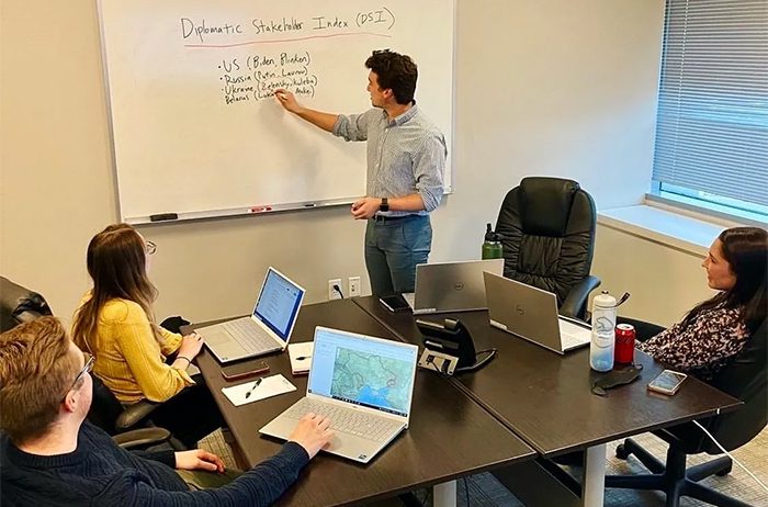 Man explaining a financial formula on a whiteboard to two colleagues in a meeting room.
