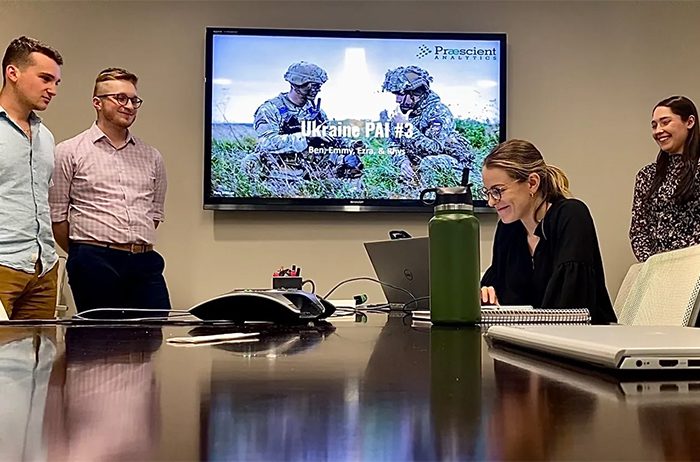 Two colleagues in a meeting room with a military presentation on screen.